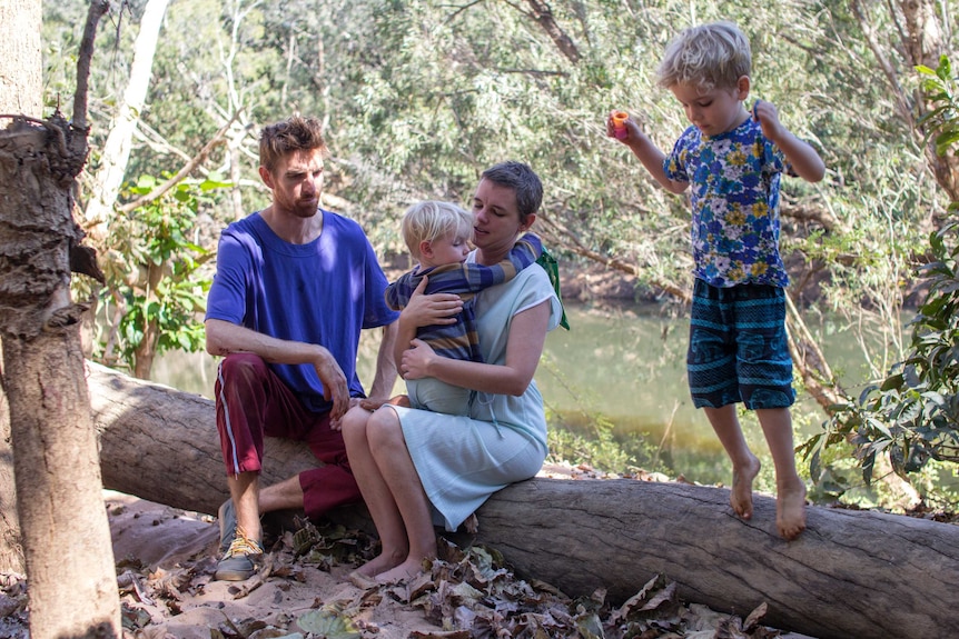 a man, a woman holding her child and a young boy on a log near a river.