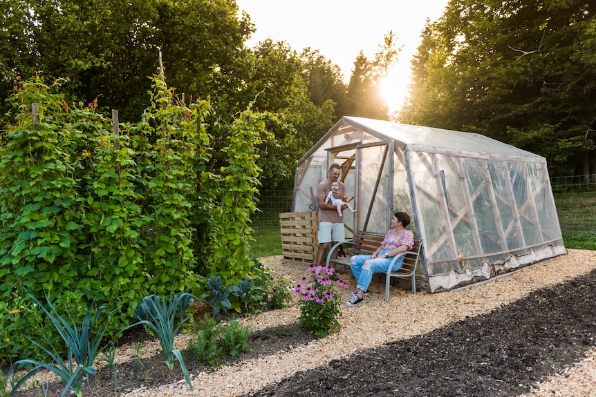 Eva and Aljaž and their baby girl stand outside their greenhouse as the sun sets behind them
