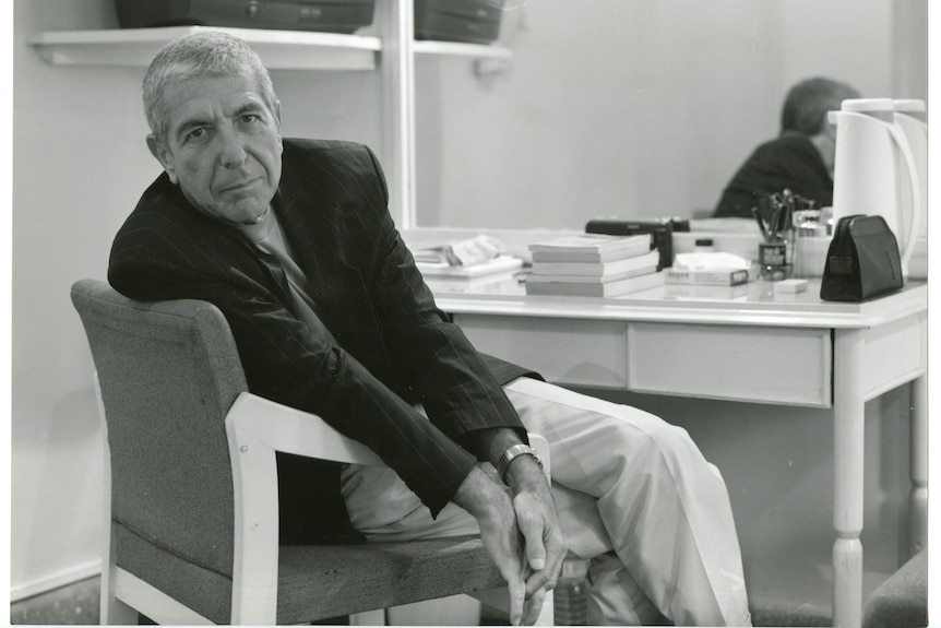 An older man sitting at a desk that is stacked with books. He is turned in his seat to look casually at the camera