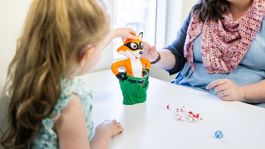 A young girl sits across a table from a woman, playing with a toy.