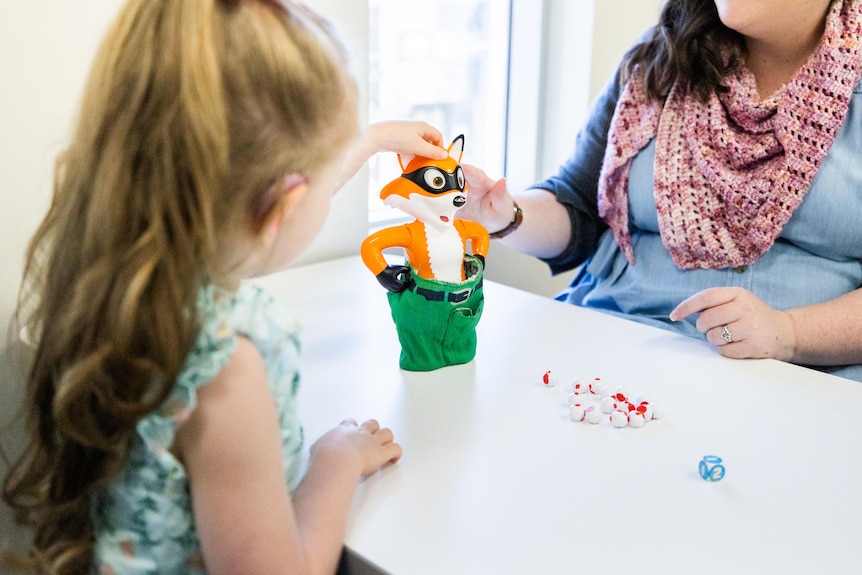 A young girl sits across a table from a woman, playing with a toy.