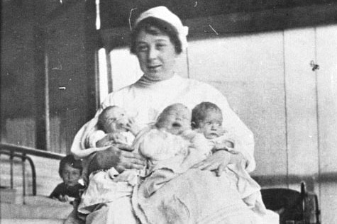 A nurse in a 1919 uniform with three babies in her arms and a small child peeking out from behind her