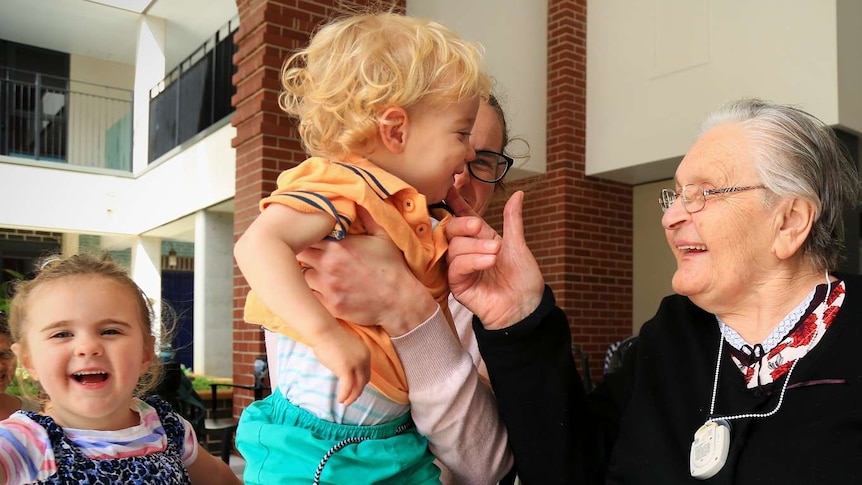 Picture of an elderly woman touching the cheek of a one year old boy who is smiling in the arms of his mother.