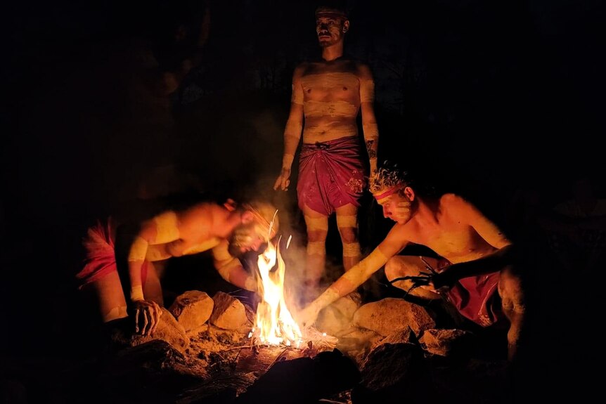 Dimly-lit wide shot of three young Aboriginal men in traditional dress lighting a fire