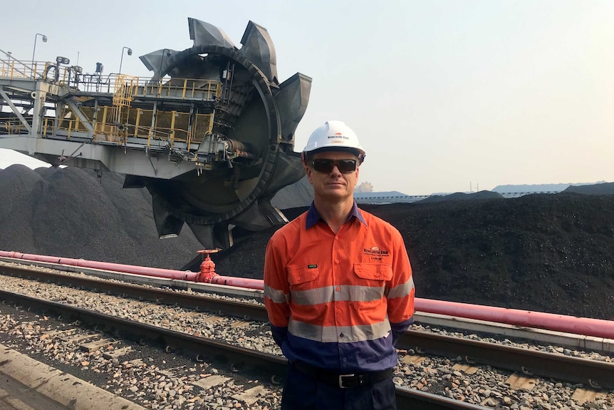 A man wearing a hi-vis shirt and hard hat stands in front of a large coal stockpile and a stacker reclaimer machine.