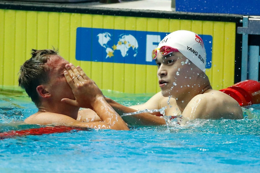 Two men hold red pool lane markers in the water near the edge of the pool's yellow wall.