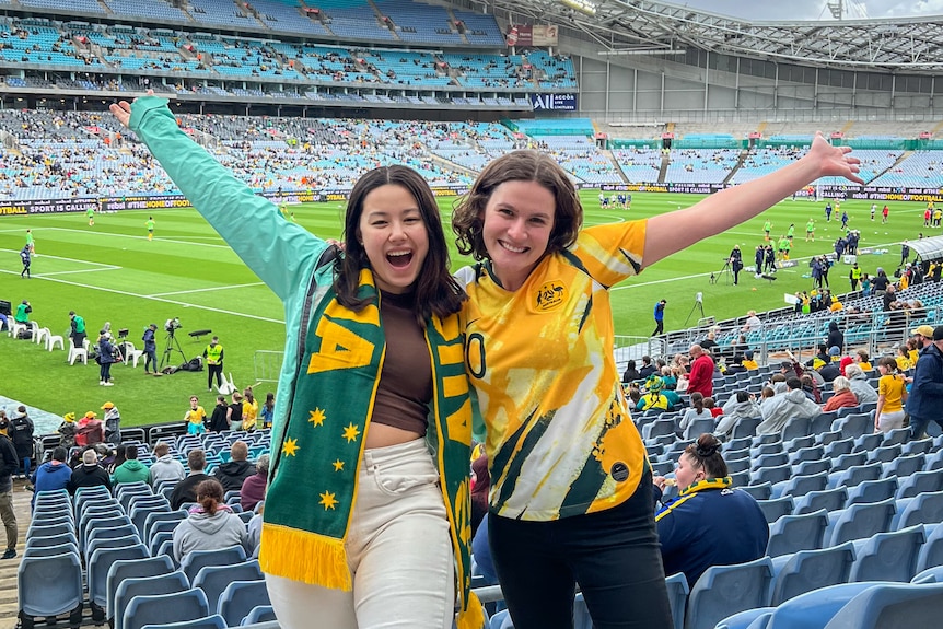 Journalist Rachel Rasker, who's wearing a Matildas jersey, grins with a friend wearing an Australia scarf in a football stadium.