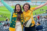Journalist Rachel Rasker, who's wearing a Matildas jersey, grins with a friend wearing an Australia scarf in a football stadium.