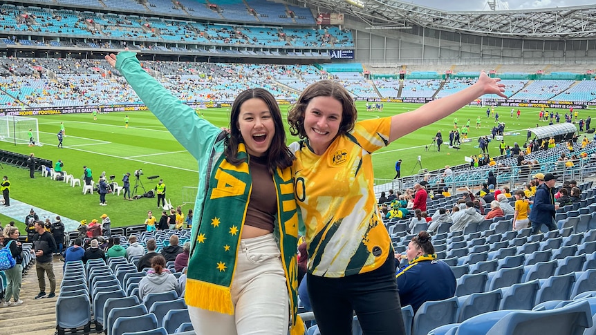 Journalist Rachel Rasker, who's wearing a Matildas jersey, grins with a friend wearing an Australia scarf in a football stadium.