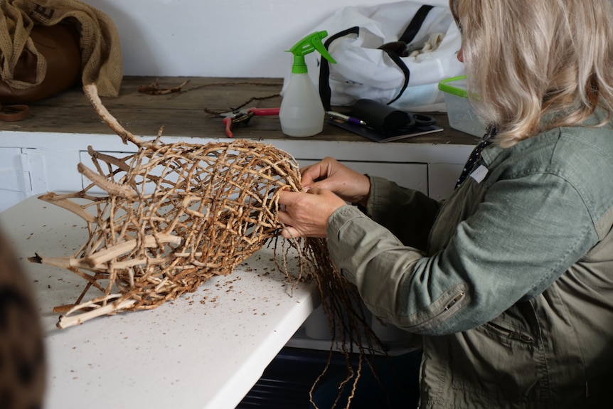 A lady sits at a table weaving a basket from brown branches.