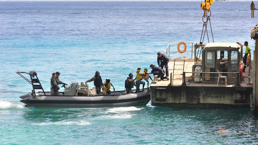 Military personnel passing a child from an inflatable boat to a barge tied to a wharf