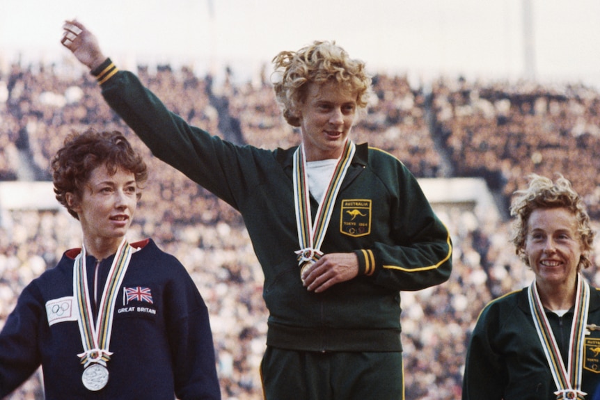 Ann Packer, Betty Cuthbert and Judith Amoore stand on the winners podium with medals around their necks.