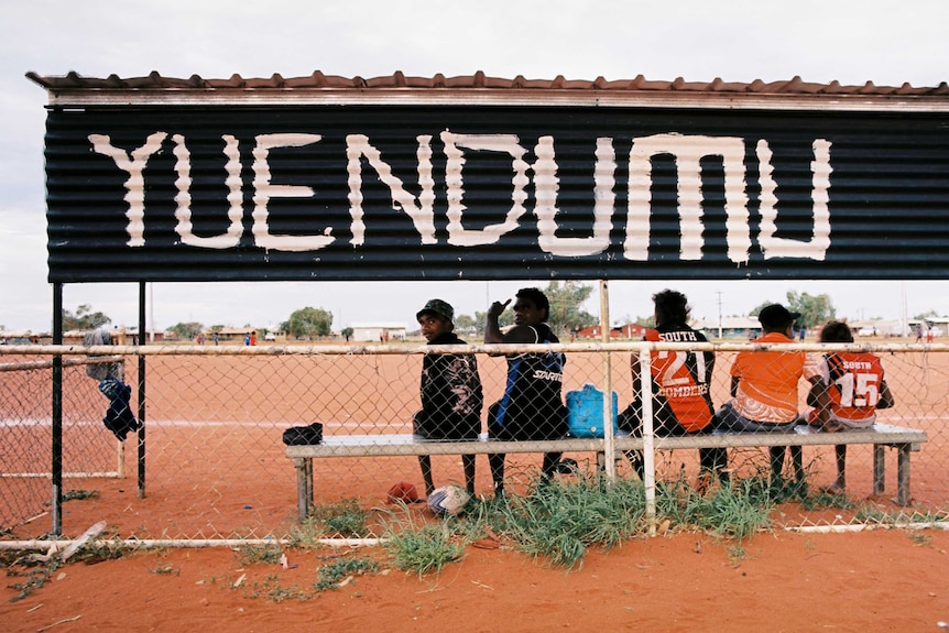 Five Indigenous youth watching football under a large sign that reads 'Yuendumu'.