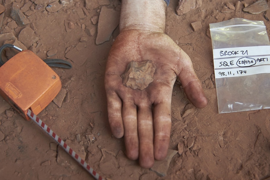 A hand holds out a rock, in the background is the dust on the Juukan rock cave floor.