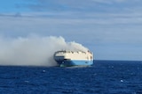 a white and blue ship can be seen from a distance at sea in the daylight with smoking billowing out of it
