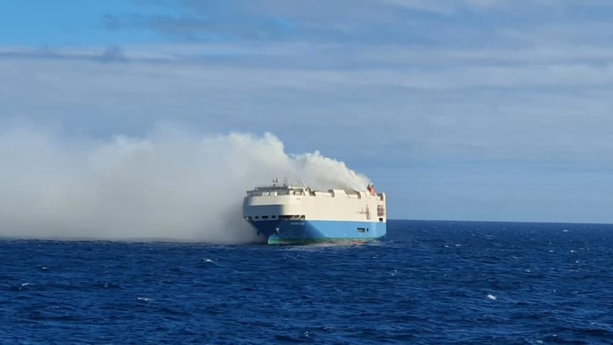 a white and blue ship can be seen from a distance at sea in the daylight with smoking billowing out of it