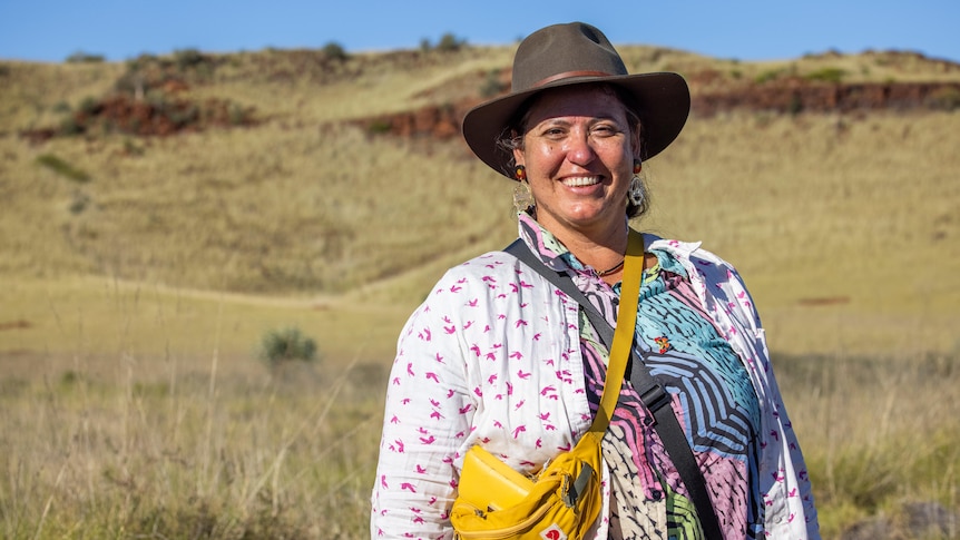 A young woman in a wide-brimmed hat smiles for a photo, with an outback landscape behind her.
