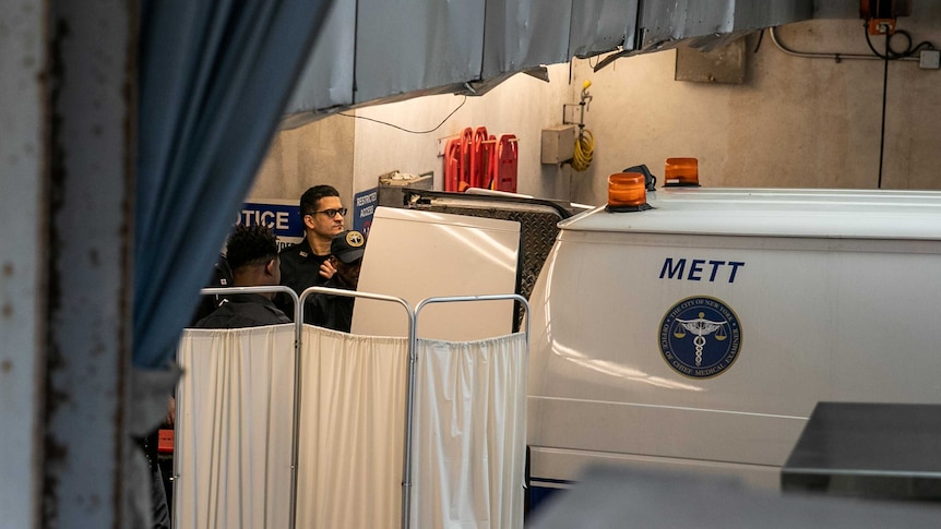 Police and medical examiners behind a van with its back doors open, behind a white sheet.