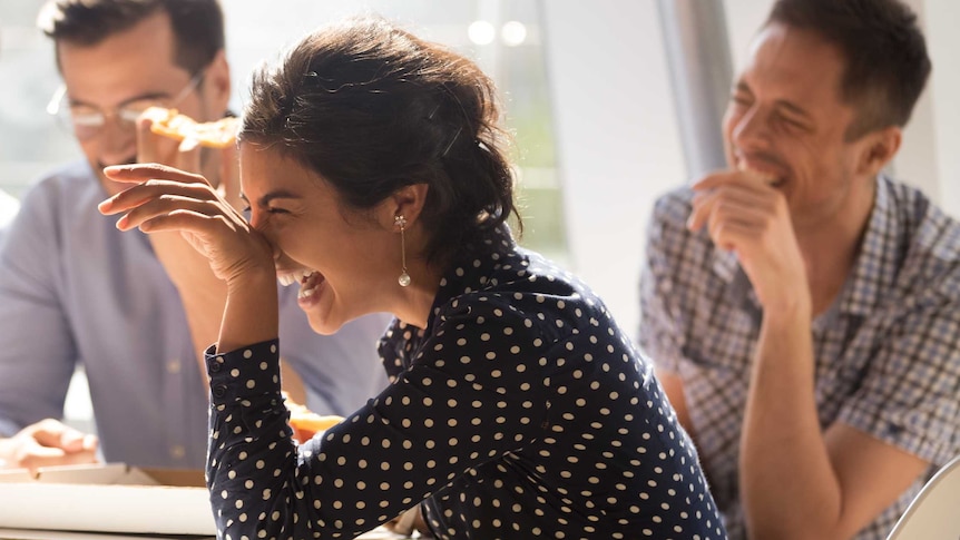 A woman dressed in spotted shirt laughs with hand at face as several others at a table smile in background.