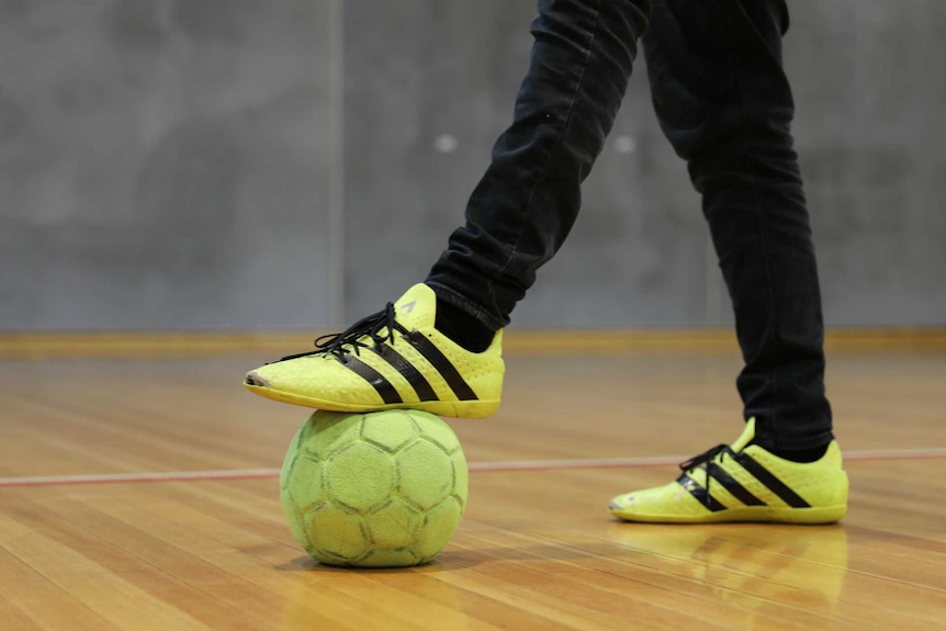 A man puts his foot on an indoor soccer ball. He is standing on a hardwood indoor court.