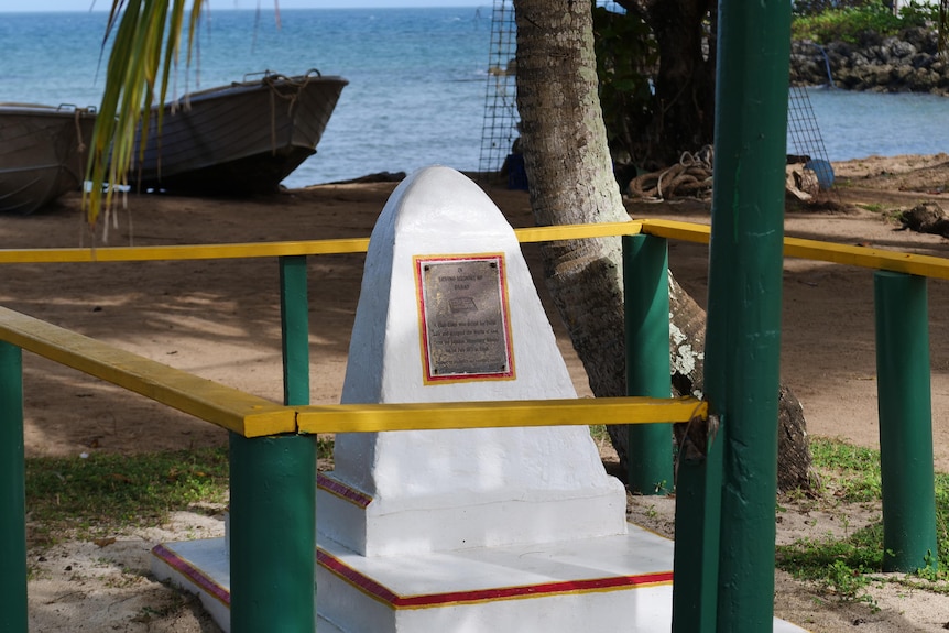 A small white monument fenced off with yellow and green wood, with water and boat in the background.