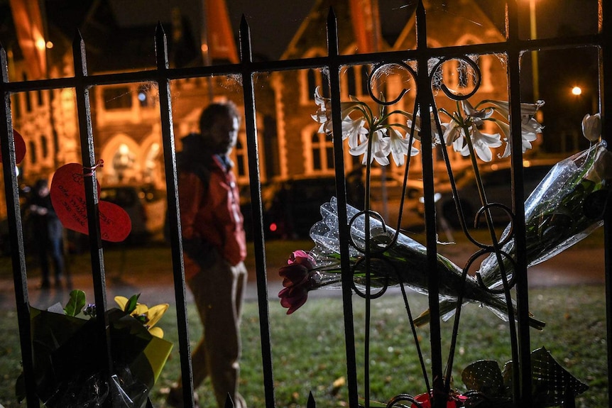 At night flowers and cards are attached to an iron gate as a man walks past.