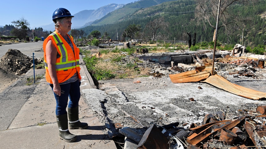 A middle-aged woman wearing a blue hard hat and orange hi-vis vest looks at burnt debris on a small concrete housing block.