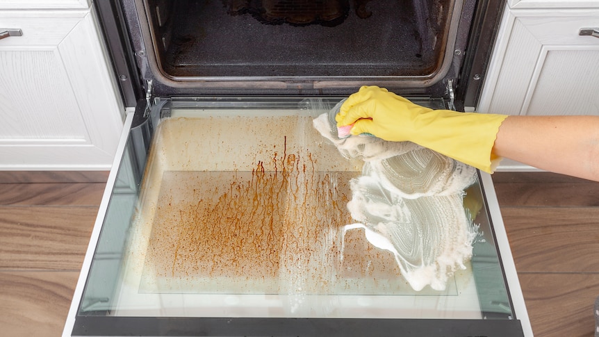 An open grimy, dirty glass oven door being scrubbed by a yellow gloved arm. 