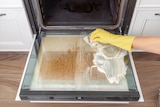 An open grimy, dirty glass oven door being scrubbed by a yellow gloved arm. 