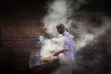 A man cooks meat over a smoky barbecue outside.