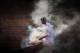 A man cooks meat over a smoky barbecue outside.