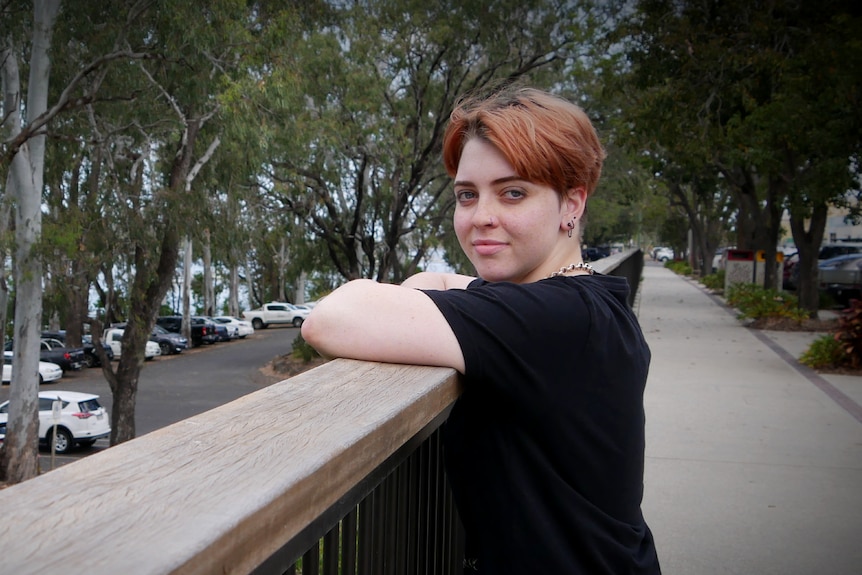 A young woman with short copper hair leaning on a fence post. She is wearing a black shirt
