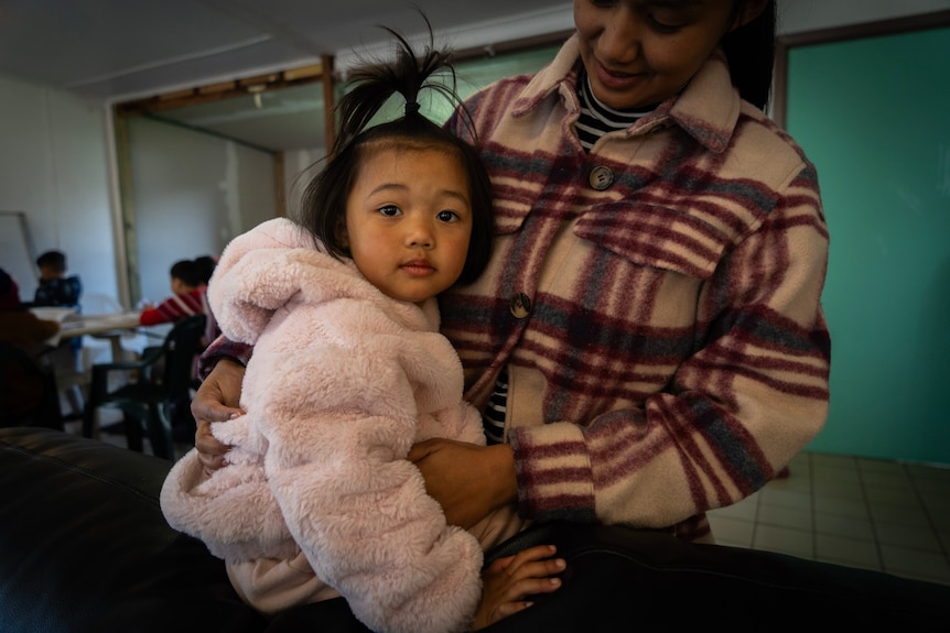 A young girl in pink hoodie cuddles is held by an adult.