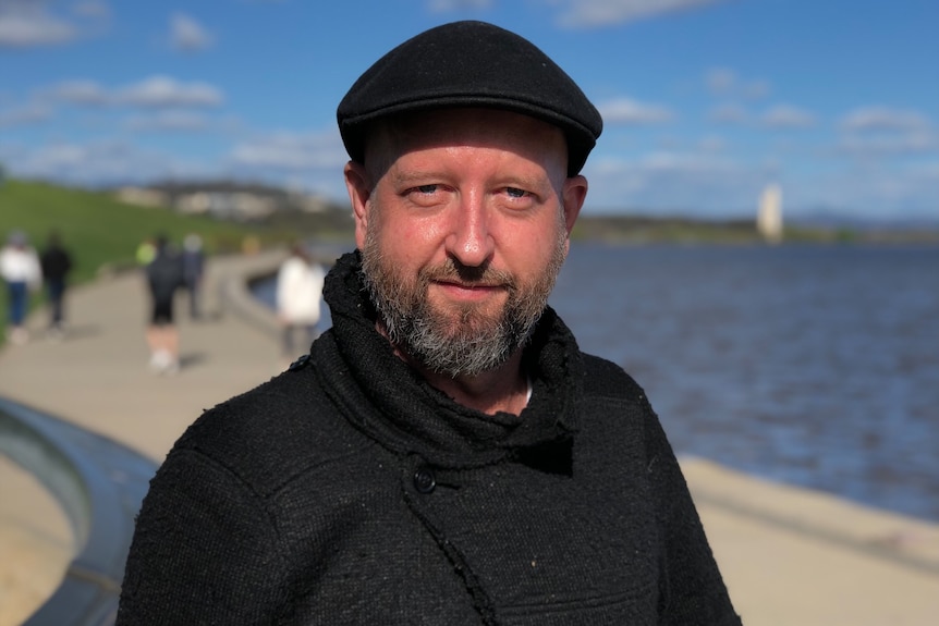 A man in a black hat stands in front of a lake.