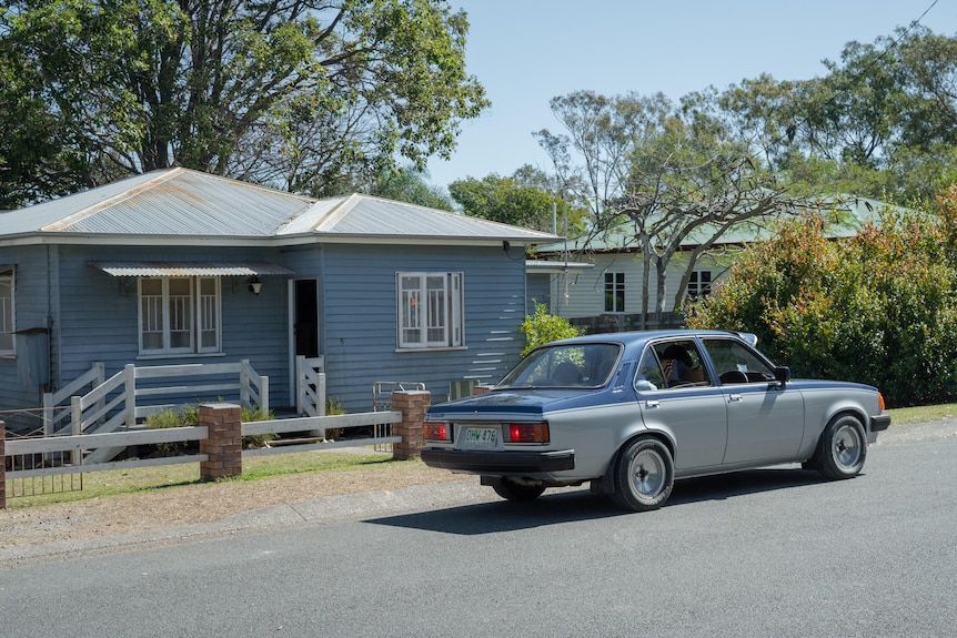 A blue house is seen with an old car parked in front of it in a quiet Queensland street on a sunny day.