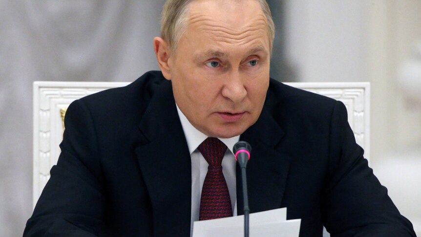 Vladimir Putin wearing a suit with a dark red tie leans over a microphone.