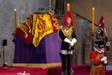 queen's coffin on a raised red platform, draped with a flag, with a guard with heads bowed