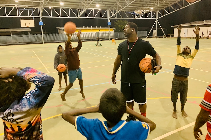A tall man stands in a group of children with basketballs on a court at nighttime 