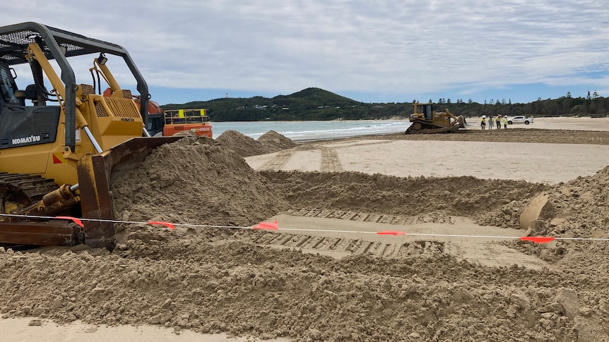 A yellow bulldozer on a beach with Cape Byron in the background.