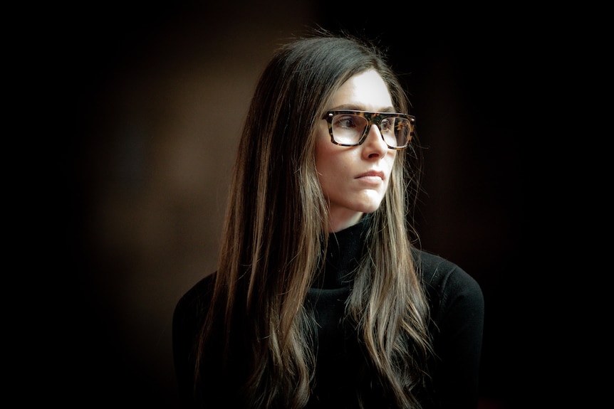 A woman with long hair, wearing a black top and dark framed spectacles, looks to her left