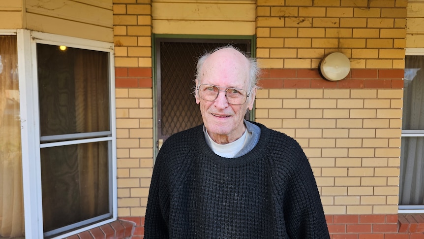 A elderly man standing in front of a brick wall wearing a sweater, he has a balding head with glasses.
