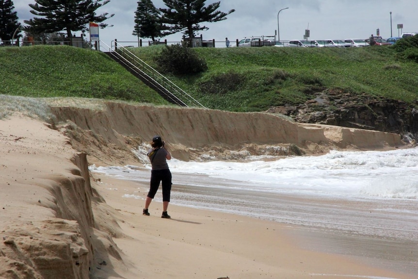 A woman stands taking pictures on a heavily eroded beach 