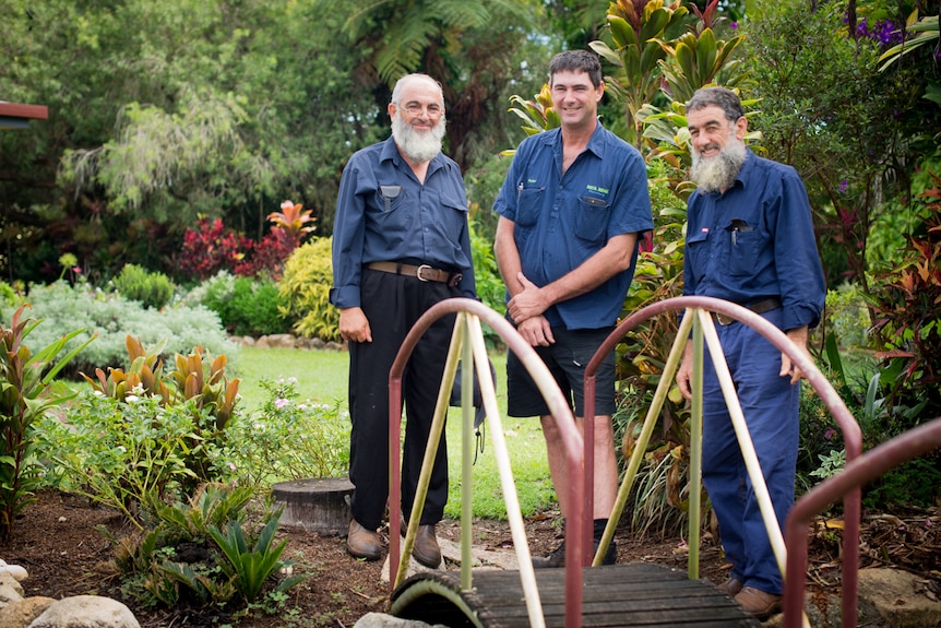 Three farmers stand next to a bridge in lush gardens.