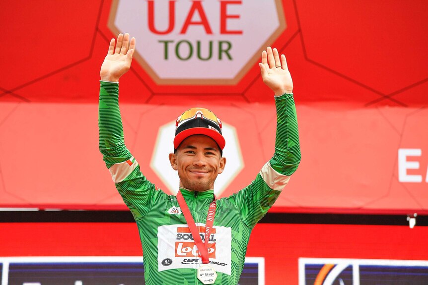 Caleb Ewan smiles and holds his arms up while wearing the green jersey and a medal around his neck.