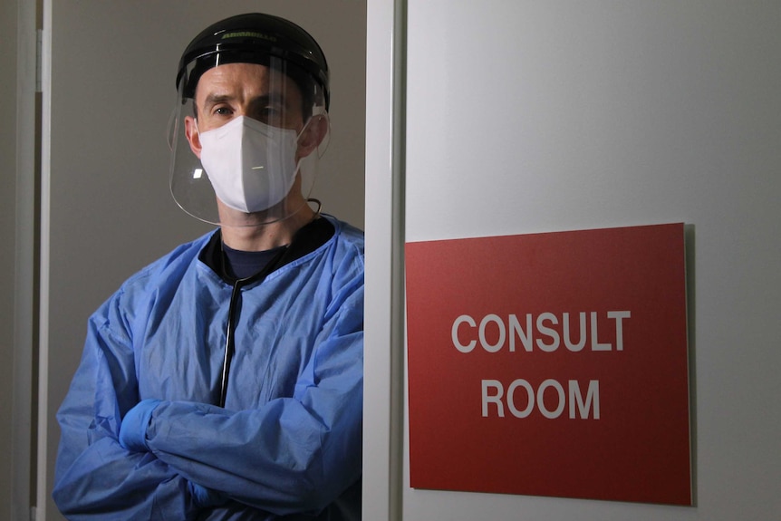 Dr McDonnell standing in full personal protective equipment in the door way, with a red sign on wall saying "consult room"