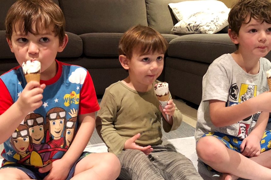 Three young boys sit eating ice creams. Two are holding them in their right hands, one is holding it in his left. 