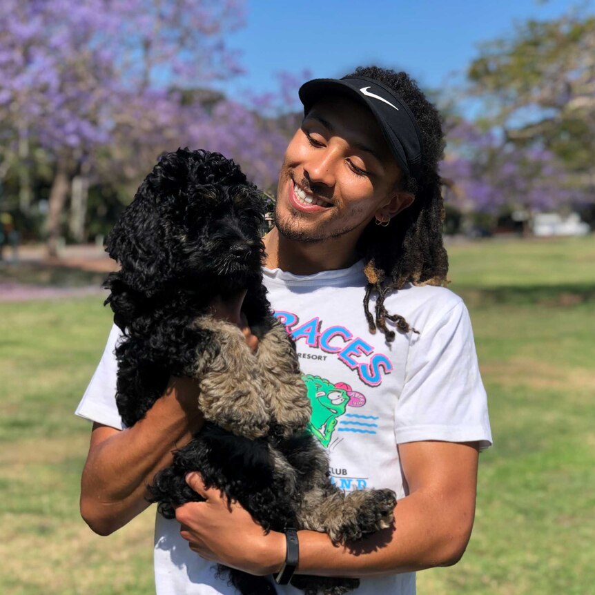 Tyler Mudge wears a white t-shirt and is in front of jacaranda trees as he holds a black puppy.
