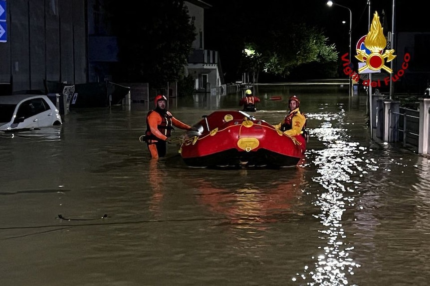 Two rescue workers ride a red dinghy boat on a flooded street at night. 