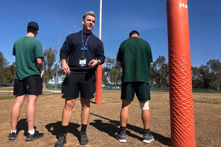 Man in black athletic gear stands next to two prisoners on dry football field