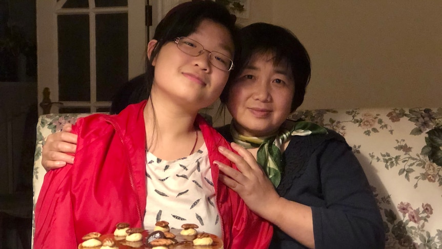 A mother and a daughter sitting in front of cake, cuddling each other.